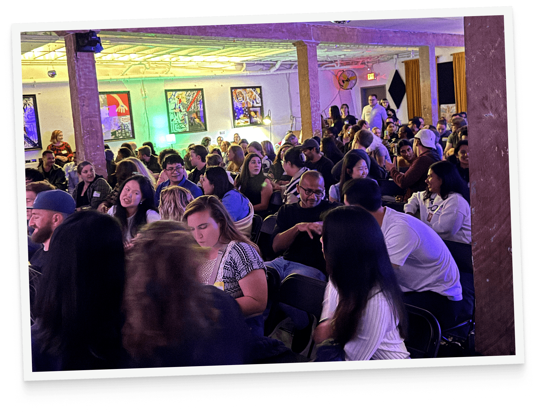 A tilted photo of a dense Big Brain crowd seated shoulder to shoulder under purple and green lighting in a warehouse venue